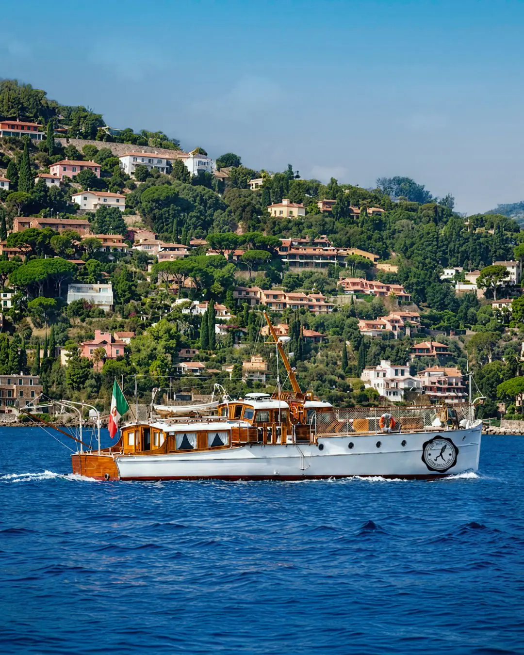 Il Katya 1929 in navigazione con la bandiera italiana, davanti a una costa verde punteggiata di ville